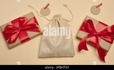 A burlap bag with ties, surrounded by two craft paper gift boxes with red bows. On a beige background Stock Photo