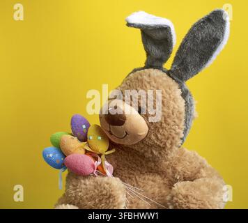Big cute brown teddy bear holding colorful Easter eggs, wearing a rabbit mask with long ears on his head, funny holiday card, close up Stock Photo