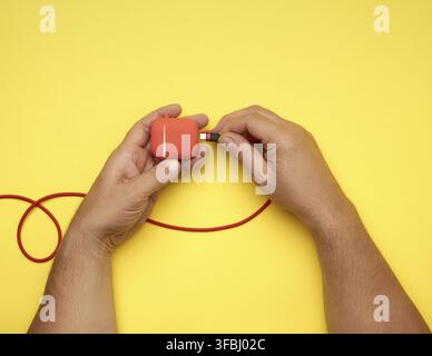 Two male hands are holding a cable and a red box with wireless headphones, yellow table, top view Stock Photo