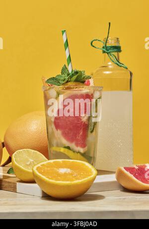 A clear glass with a cold drink, grapefruit slices, ice and mint, with a green striped straw, on a yellow background. Sliced ??citrus fruits lie nearb Stock Photo