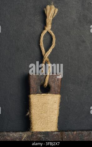 An old cutting board handle made of dark wood, wrapped in natural jute twine, with a loop at the top. It rests on a black textured background Stock Photo