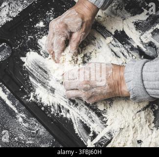 Two female hands knead the dough from white wheat flour on a black table, top view Stock Photo