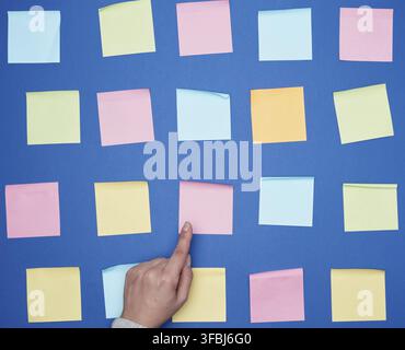 Female hand and a lot of empty paper multi-colored square stickers on a blue background, concept of choosing a strategy with the specified finger Stock Photo