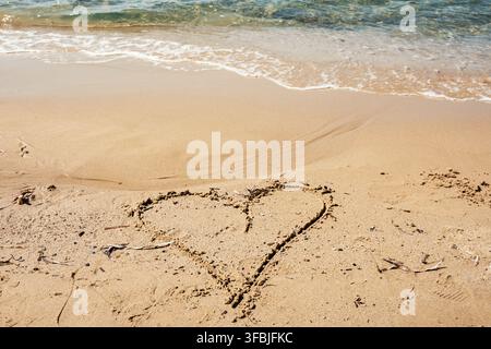 Hand drawn heart symbol in the golden sand with gentle sea waves washing ashore during a sunny summer vacation day in Greece. Stock Photo