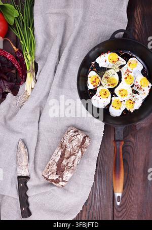 Fried quail eggs in a cast-iron black frying pan with a wooden handle on the table, next to a whole loaf of rye bread, top view Stock Photo