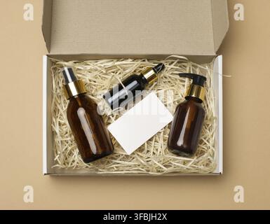 Brown glass bottles for cosmetics and a white paper blank business card lie in a cardboard paper box on a beige background, top view. Delivery Stock Photo