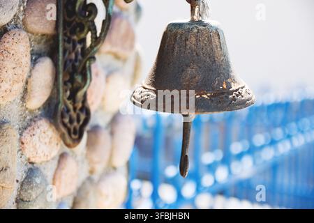 Close-up of an old rusty metal bell mounted on a textured stone wall with decorative ironwork in Greece. Stock Photo