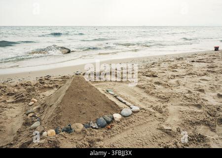 A pyramid shaped sand sculpture surrounded by small round stones on a sandy coastline with gentle sea waves washing ashore, Greece. Stock Photo
