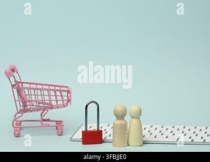 A red metal padlock stands on the keyboard and behind a miniature shopping cart, blue background. Personal data protection concept, secure online shop Stock Photo