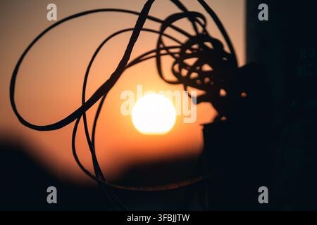 Tangled electric cables frame the bright sunset sky with a warm golden glow. A close up view of messy wires silhouetted against the glowing evening su Stock Photo
