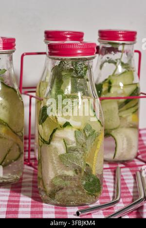 Glass bottles with red lids, filled with detox water containing cucumber and lemon slices and mint leaves, on a red and white checkered tablecloth. Me Stock Photo