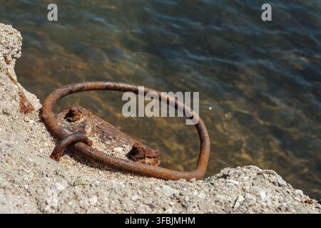 Rusty metal mooring ring attached to weathered concrete structure by the sea with clear water ripples. Greece. Stock Photo