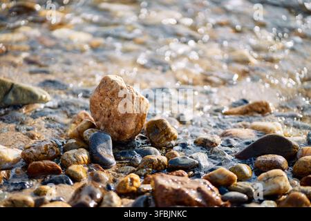 Close-up view of smooth multi-colored pebbles and wet sand on a sea shore with sparkling water ripples washing over the ground in Greece. Stock Photo