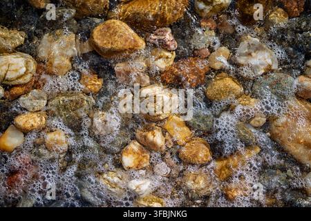 High angle close-up shot capturing wet colorful pebbles and sparkling sea foam washing over a rocky beach shore. Stock Photo