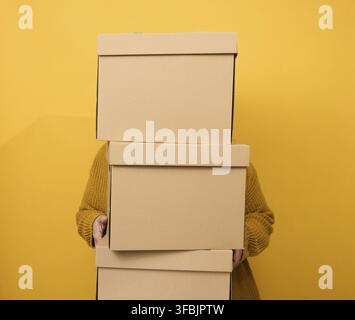 A woman in a sweater holds a stack of brown cardboard boxes on a yellow background. Relocation Stock Photo