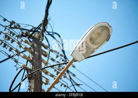 An old street light fixture mounted on a weathered wooden utility pole surrounded by a complex network of tangled power lines and cables against a cle Stock Photo