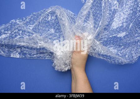 Female hand holding crumpled piece of polyethylene with air bubbles on a blue background, packing material Stock Photo