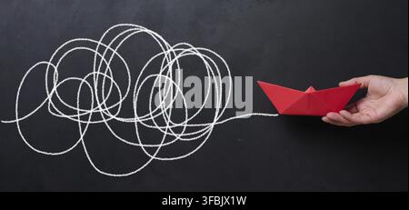A female hand holds a red paper boat against a black chalkboard background with a long path. Concept of mentorship and assistance Stock Photo