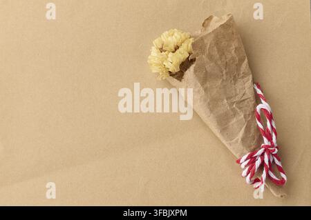 A small bouquet of yellow dried flowers in a craft paper bag tied with red and white twine on a brown background Stock Photo