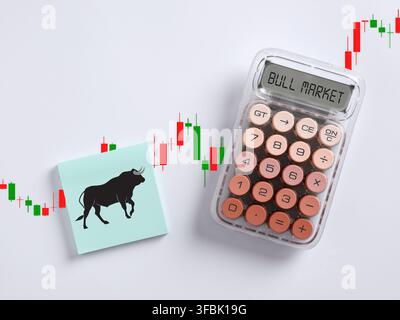 Bull market concept showing a calculator displaying BULL MARKET and a sticky note with a bull icon on a financial candlestick chart background Stock Photo