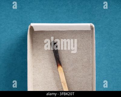 Single burnt matchstick resting inside an open cardboard box against a blue background, representing the end of a process Stock Photo
