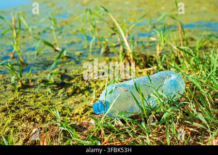 A discarded clear plastic water bottle lying in green grass along an algae-covered shoreline of a river. Selective focus. Stock Photo