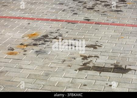 Dirty dark motor oil stains and rust marks spill on gray cobblestone sidewalk pavement outdoors in an urban parking lot on a bright sunny day. Stock Photo