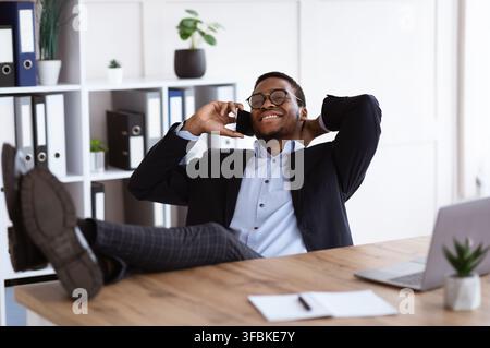 Relaxed black manager sitting with legs on desk, having conversation Stock Photo