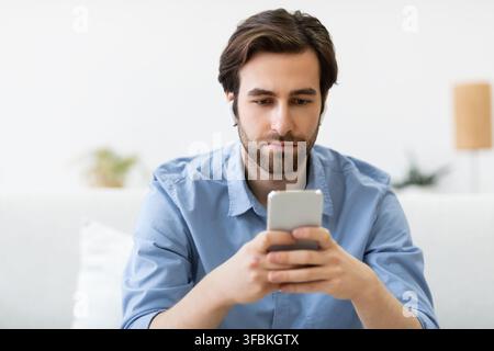Serious Man Using Smartphone Wearing Earbuds Sitting On Sofa Indoors Stock Photo