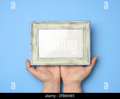 Two female hands holding a rectangular blank wooden frame, blue background Stock Photo