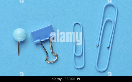 Blue stationery including a pushpin, a binder and two paper clips of different sizes arranged in a row on a blue background Stock Photo