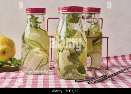 Glass bottles with red lids, filled with detox water containing cucumber and lemon slices and mint leaves, on a red and white checkered tablecloth. Me Stock Photo