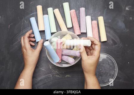 Multicolored chalk in a plastic bucket on a black background, female hands fold objects into a container, top view Stock Photo