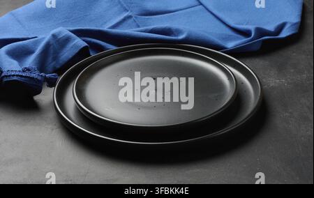 Empty black ceramic plates stacked on dark background with blue linen napkin. Minimalist food still life Stock Photo