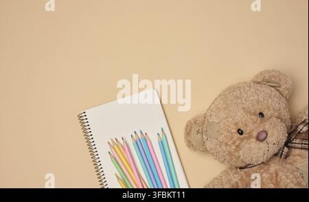 Wooden colored pencils and open notebook, brown teddy bear next to it on beige background, top view Stock Photo