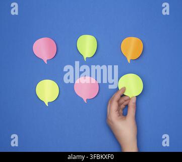 Empty paper multi-colored stickers in the shape of a cloud and a female hand glues one piece of paper on a blue background, dialogue concept Stock Photo