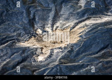 Horizontal high angle macro shot of dark blue crumpled plastic tarp surface covered with light sand and dust spot in bright daylight. Stock Photo