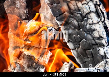 Horizontal close up macro shot of hot orange flames glowing between charred black wood logs and grey ashes in an outdoor fire pit without people. Stock Photo