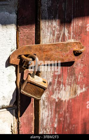 Vertical view of an old unlocked rusty padlock on a metal latch mounted on a weathered red painted wooden door next to a white wall. Stock Photo