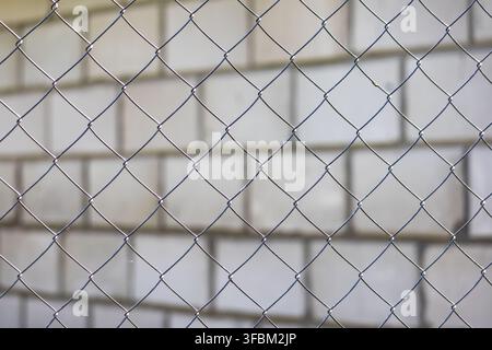 Close up horizontal view of gray metal chain link fence net with a blurred light brick wall texture behind it on a sunny day. Stock Photo