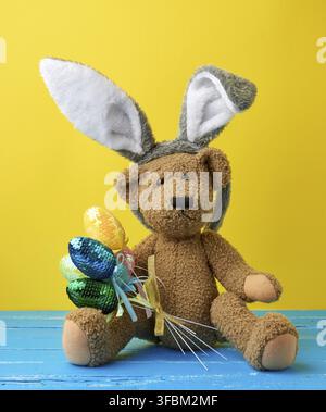 Vintage cute brown teddy bear holding colorful Easter eggs, wearing a rabbit mask with long ears on his head, funny holiday card Stock Photo