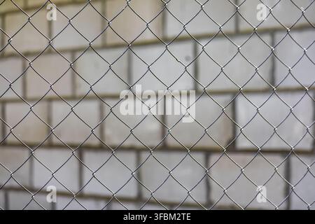 Horizontal close up view of a metallic chain link fence mesh against a soft blurred white brick wall background outdoors in daylight. Stock Photo