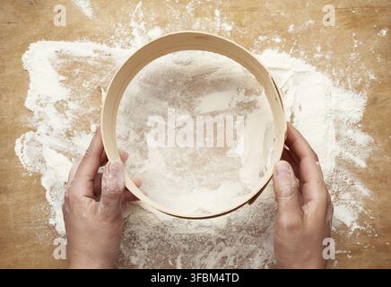 Two female hands hold a round wooden sieve over a table with white flour, top view Stock Photo