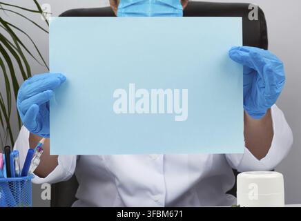 Doctor in a white medical coat is sitting at a table in a brown leather chair and holding a empty blue sheet of paper in his hands, place for an inscr Stock Photo