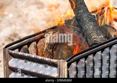 A close-up view focusing on the textured side panel and open end of a metal barbecue grill, filled with burning wood logs actively engulfed in flames Stock Photo