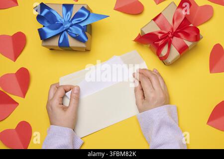 Female hands hold a white rectangular envelope on a yellow background, paper red hearts. View from above Stock Photo