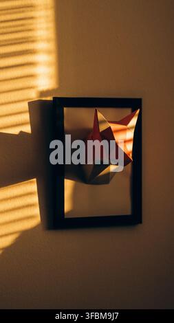 View of a low-poly paper fox head sculpture mounted in a black frame on a wall, illuminated by warm sunlight casting striped shadows in Svitavy, CZ. Stock Photo