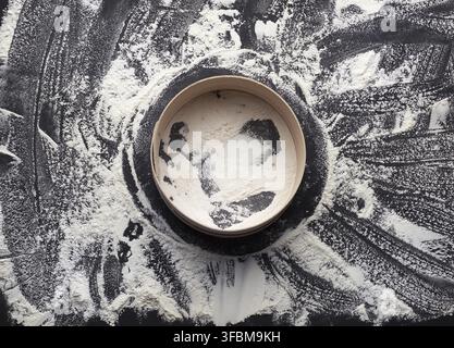 Sprinkled white wheat flour and a round wooden sieve on a black table, top view Stock Photo