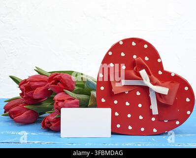 Empty rectangular white paper business card, bouquet of red tulips and a heart-shaped cardboard box on a blue wooden background. Festive backdrop for Stock Photo