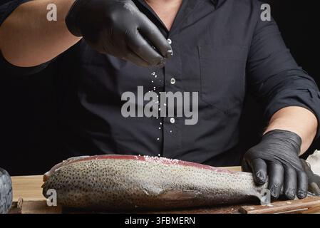 Chef in a black shirt and black latex gloves prepares salmon fillet on a wooden cutting board, process of sprinkling with spices and salt, low key Stock Photo